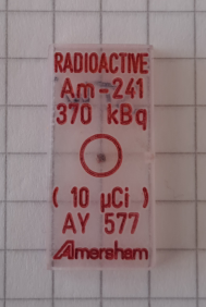 Foto des 241-Am-Punktstrahlers aus dem unten abgebildeten Set. Der Strahler (dunkler Punkt in der Mitte des roten Kreises) ist in eine rechteckige Plastikmatrix eingebettet.